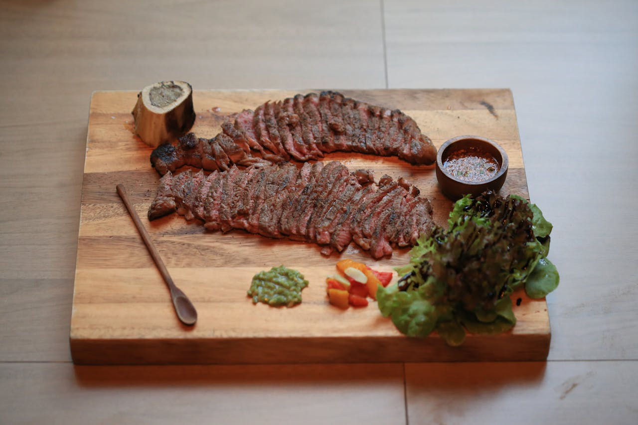 Sliced grilled steak with green pesto, sauce, and lettuce on a wooden cutting board.